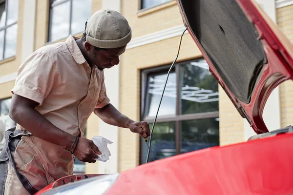 Hombre checando el nivel de aceite para transmisión automática sacando la varilla medidora debajo del capó rojo de su coche.