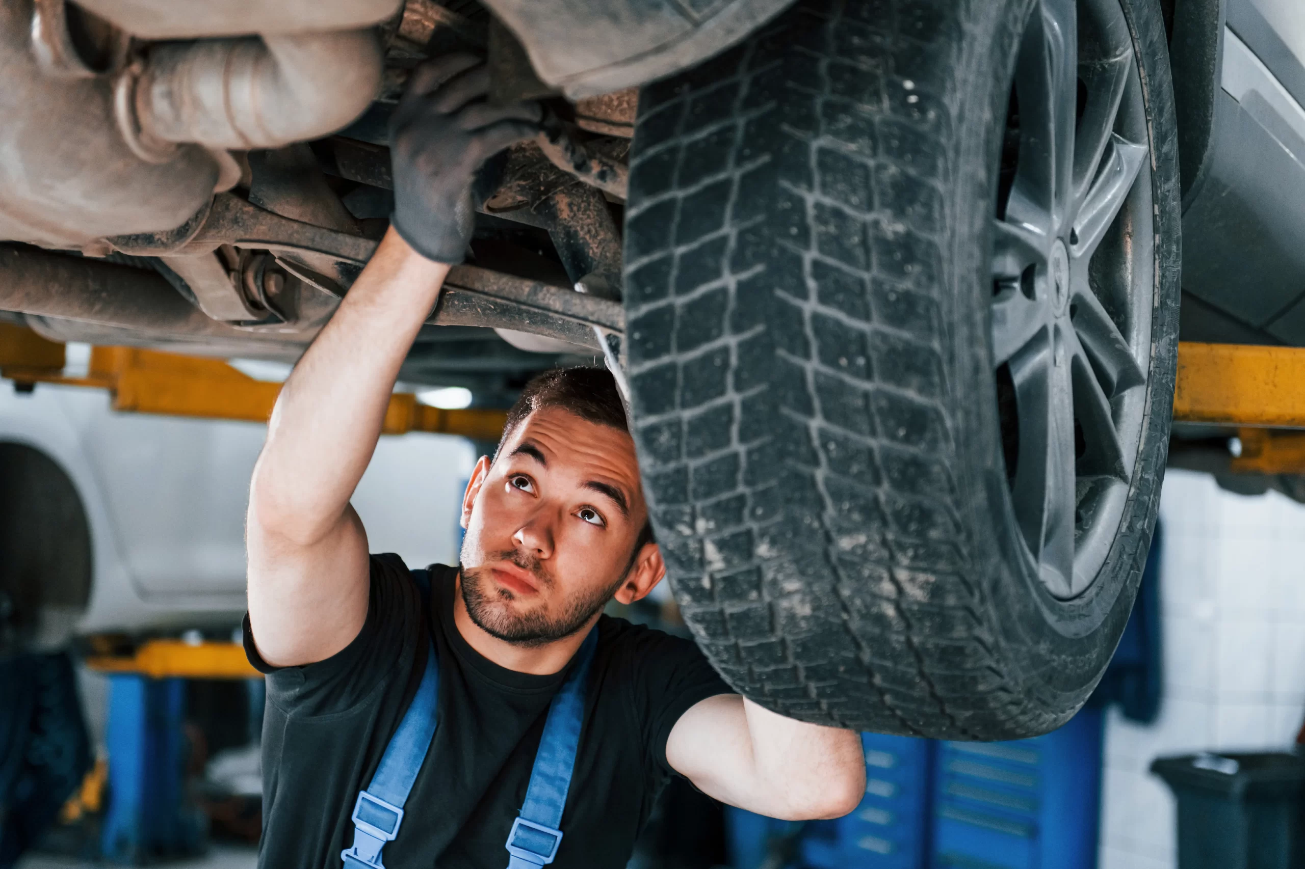Un joven mecánico con uniforme de trabajo revisa cuidadosamente la parte inferior de un coche elevado, posiblemente el chicote de cambios o la suspensión
