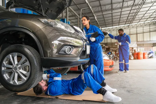 Mecánico acostado en el suelo bajo un SUV mientras una compañera lo supervisa, realizando trabajo en un taller de transmisiones automáticas.
